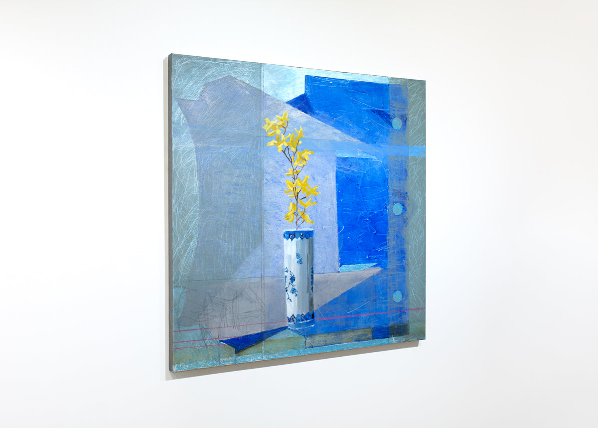 An abstracted blue painting with a vase with yellow flowers in the foreground hangs on a white wall.