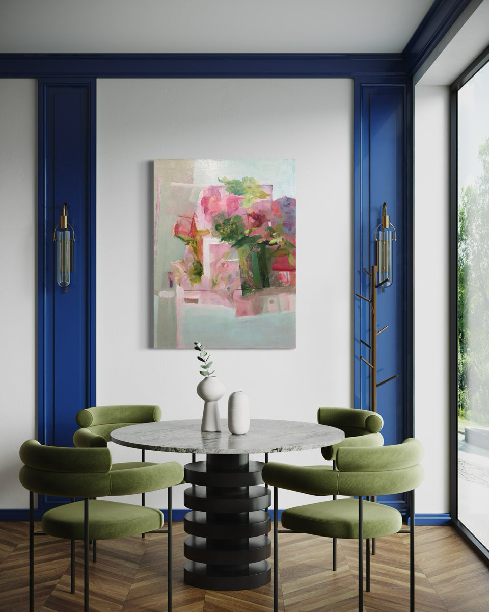 A painting depicting a green vase full of lush, pink roses and peonies is hung above a dining table with green chairs. This painting is wired and ready to hang.