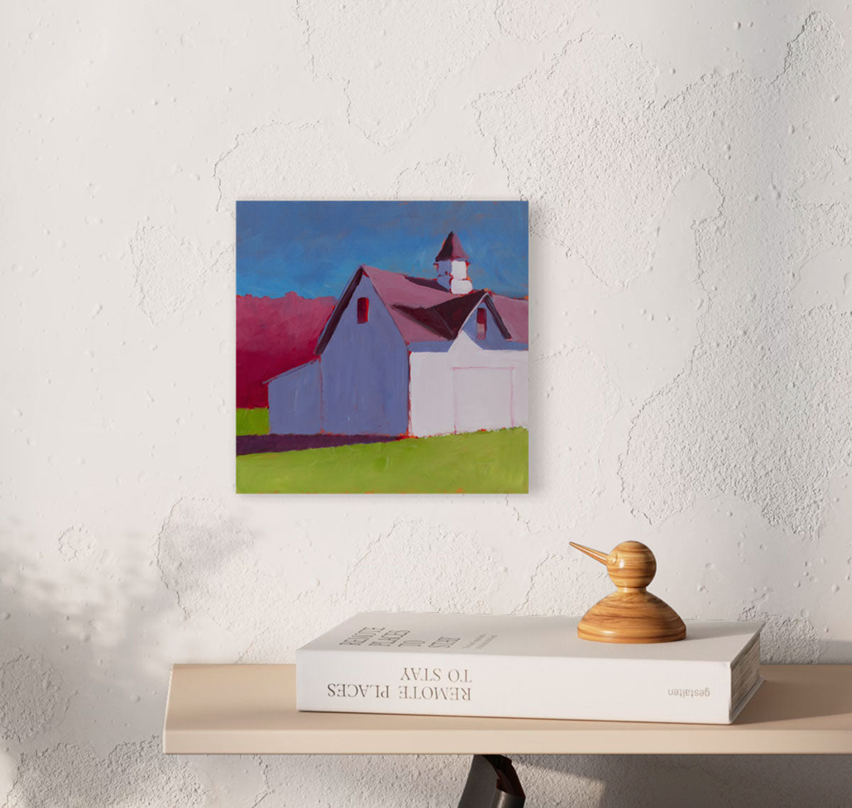A pink, blue, and green contemporary landscape painting hangs on a white textured wall above a floating wood shelf on which a white book and decor rest.