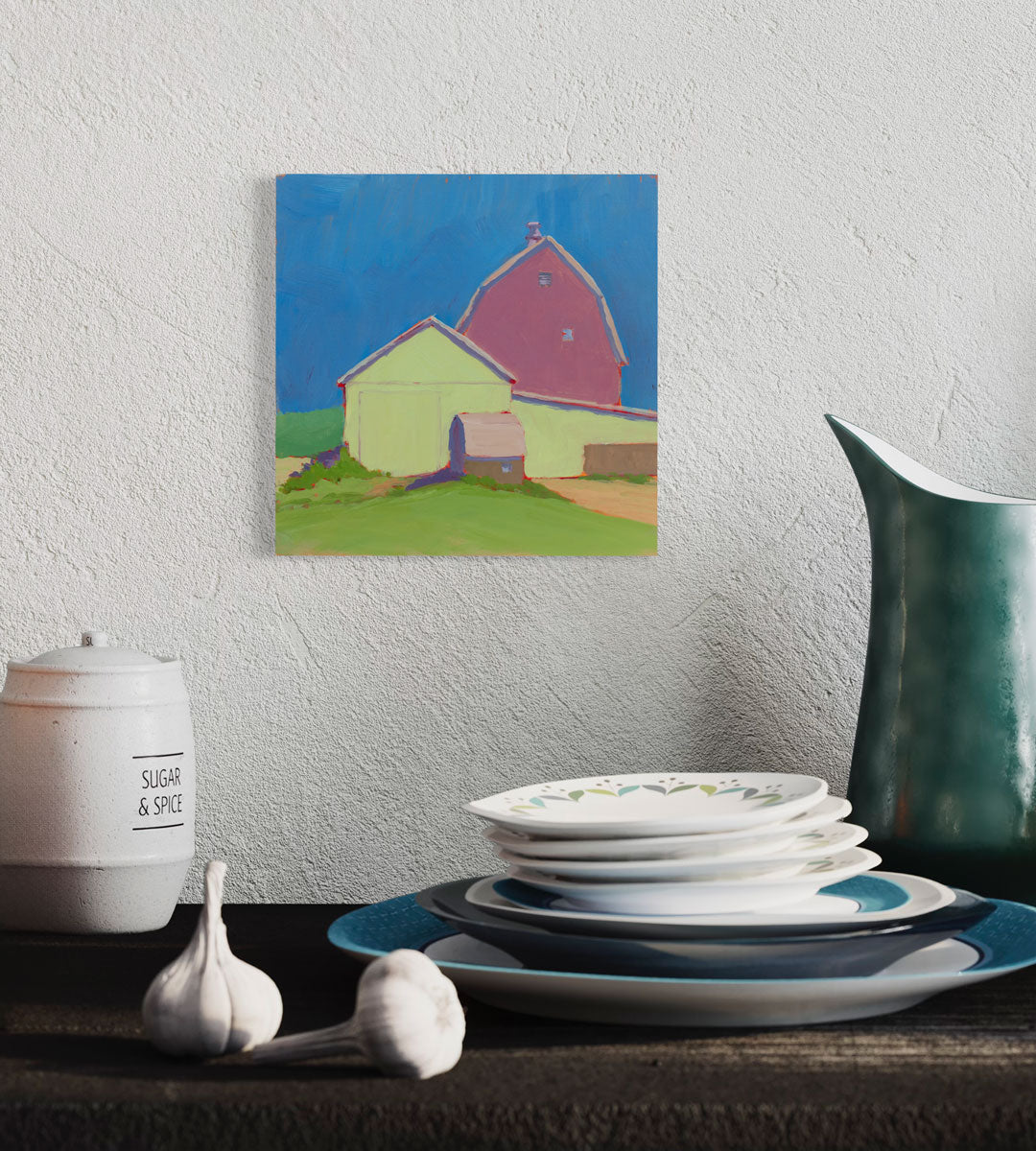 A blue, green, and pink contemporary landscape painting hangs on a textured white wall above a stack of plates and other kitchen items.