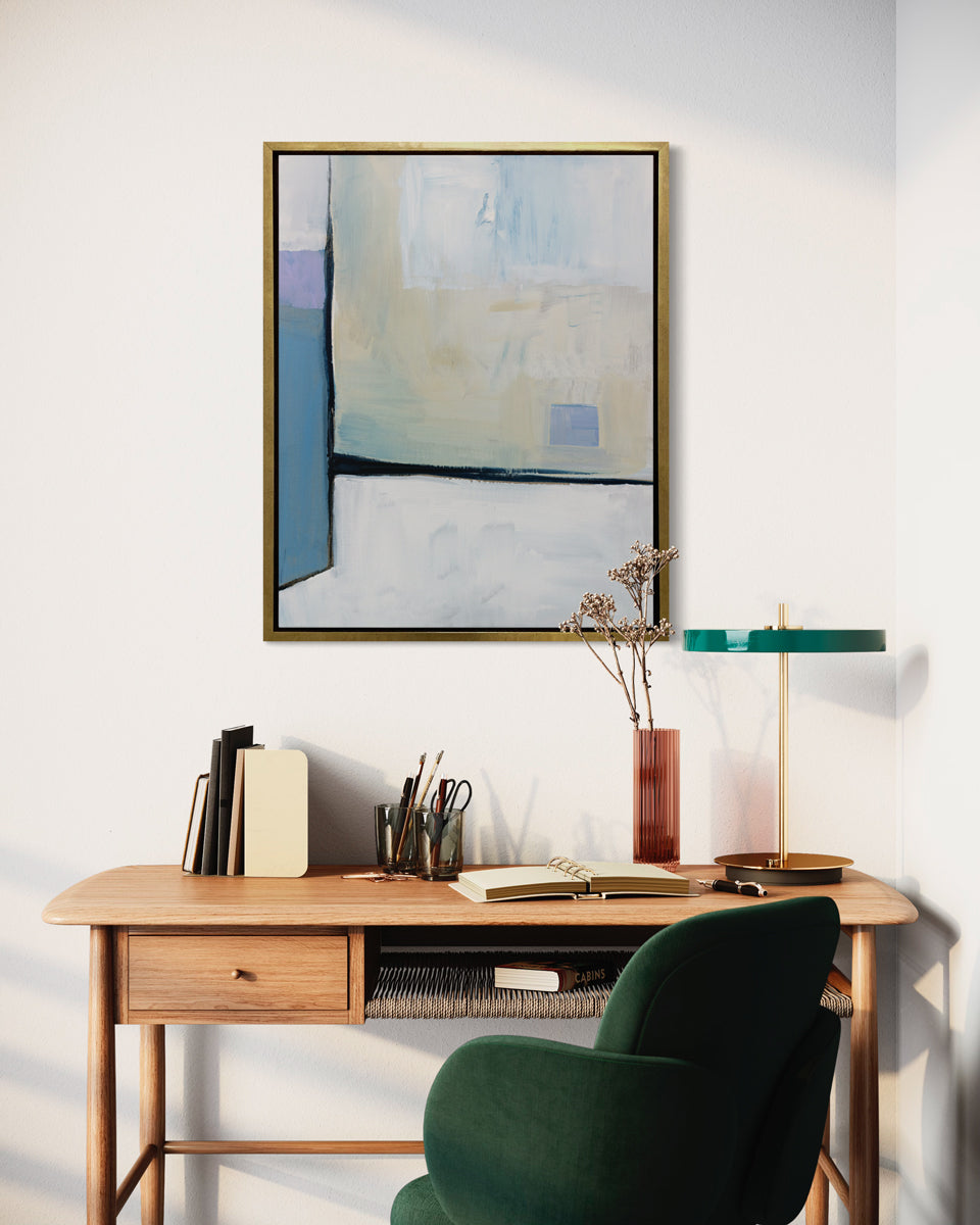 A white, black, blue and chartreuse abstract print in a gold floater frame hangs on a white wall above a natural wood desk with notebooks, a modern lamp and a deep green chair.