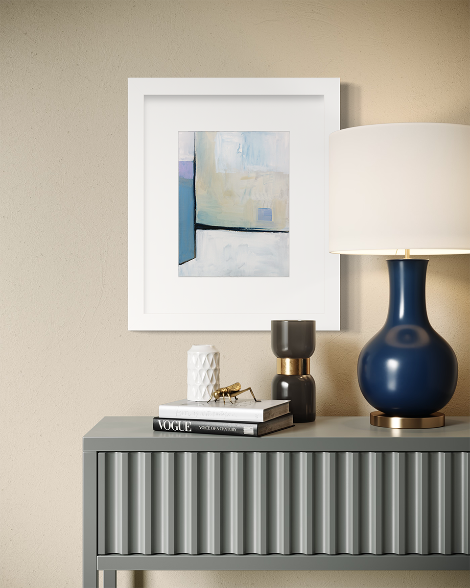 A white, black, blue and chartreuse abstract print in a white frame with a mat hangs on a beige wall above a grey console table with a blue table lamp, a stack of books and some decorative accessories.