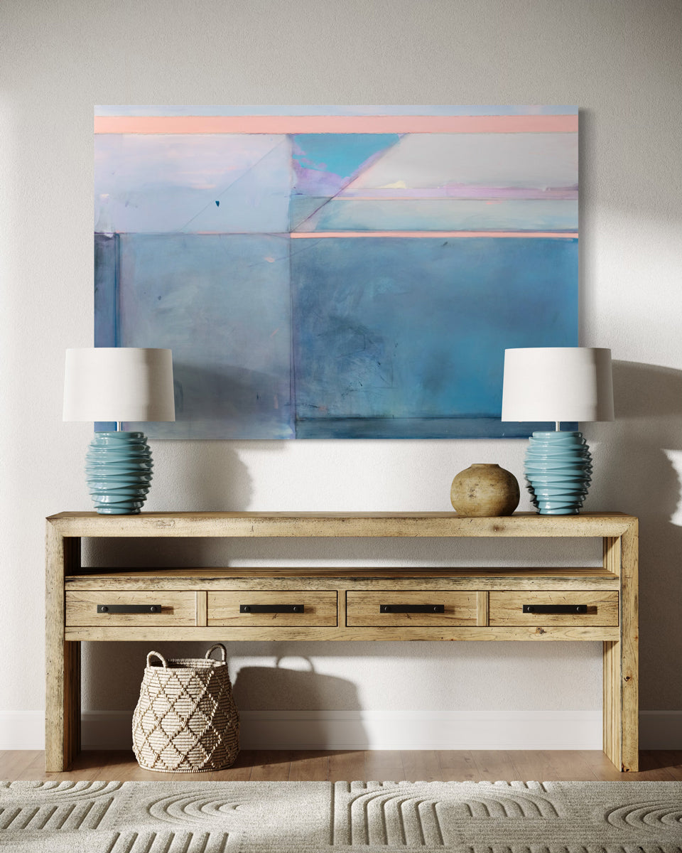 A blue and pink abstract painting hangs on a white wall. over a natural wood credenza with 2 teal ceramic lamps.