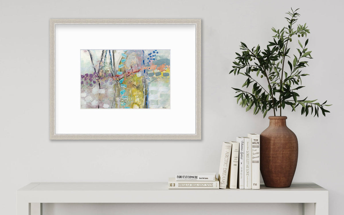 A multicolored abstract print in a silver frame with a mat hangs on a white wall above a white console table with a stack of books and a terracotta vase with greenery stems.