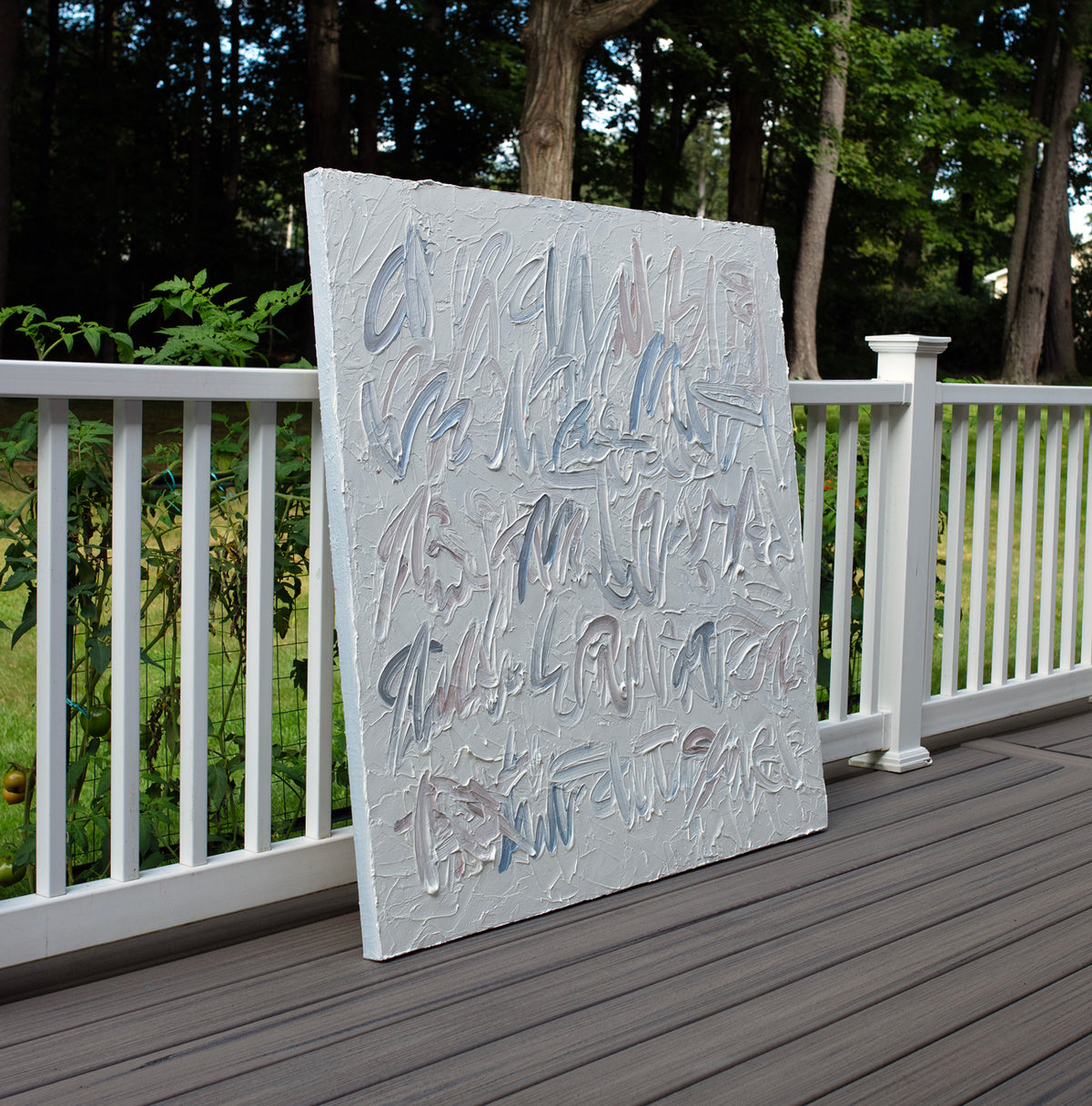 An abstract canvas text based painting with thick, textured impasto brushstrokes in shades of gray, white, and muted blue, leaning against a white wooden railing on a dark wood deck with a lush green garden background.