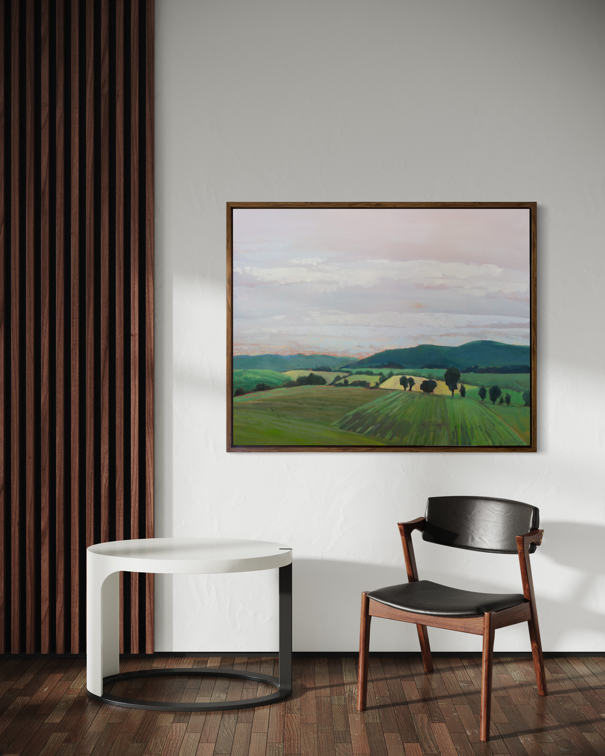 A landscape print in an oak wood floater frame hangs on a white wall above a black and oak chair and a white and black round coffee table.