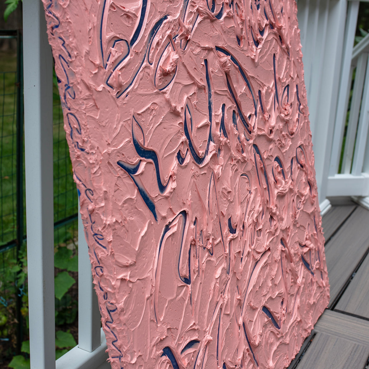 An abstract textured canvas painting leaning against a white railing on a wooden deck. The artwork features a thick, impasto pink background with dark navy blue calligraphic brushstrokes.