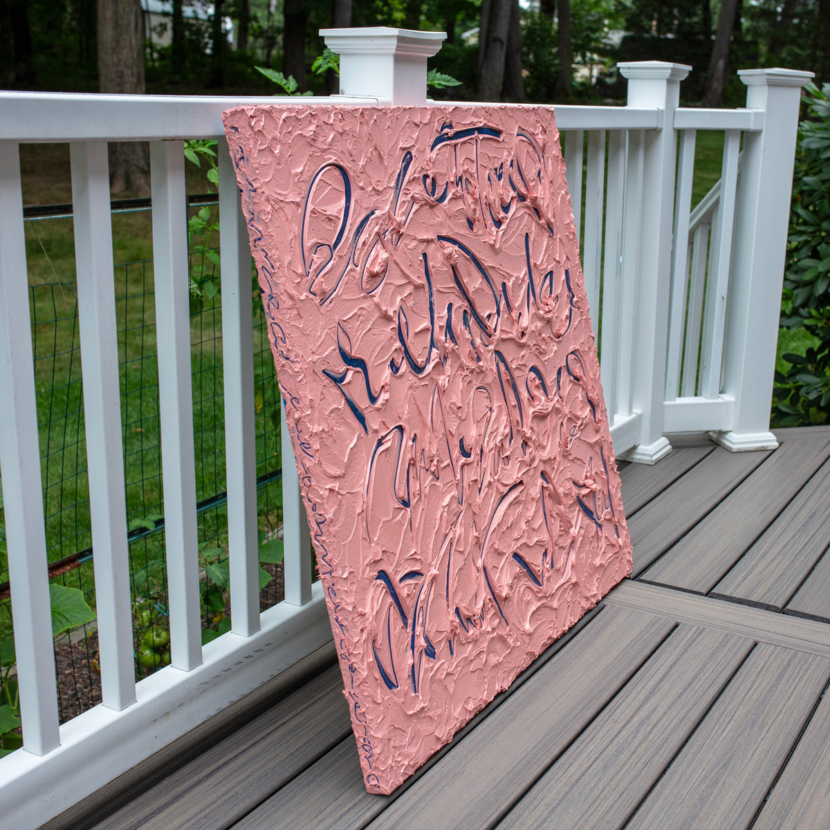 A square canvas painting with a textured pink background and dark blue abstract calligraphic brushstrokes, leaning against a white wooden deck railing with green grass and trees in the background.