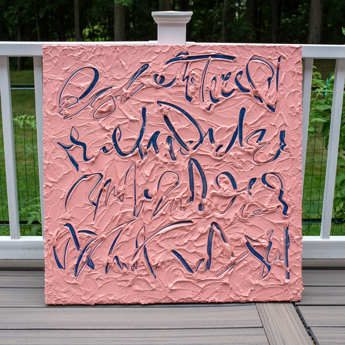 A square text based painting featuring abstract navy blue calligraphic brushstrokes over a textured pink background, displayed on a wooden deck railing with a white fence and green trees in the background.