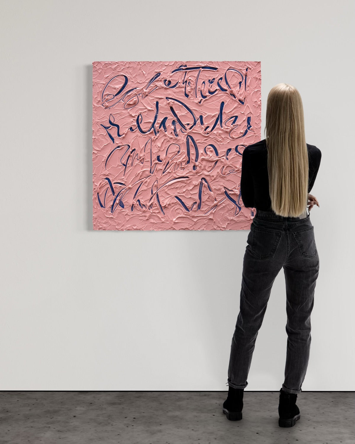 A woman with long blonde hair stands with her back to the camera, looking at a large textured pink canvas painting with dark blue calligraphic lettering mounted on a white gallery wall.