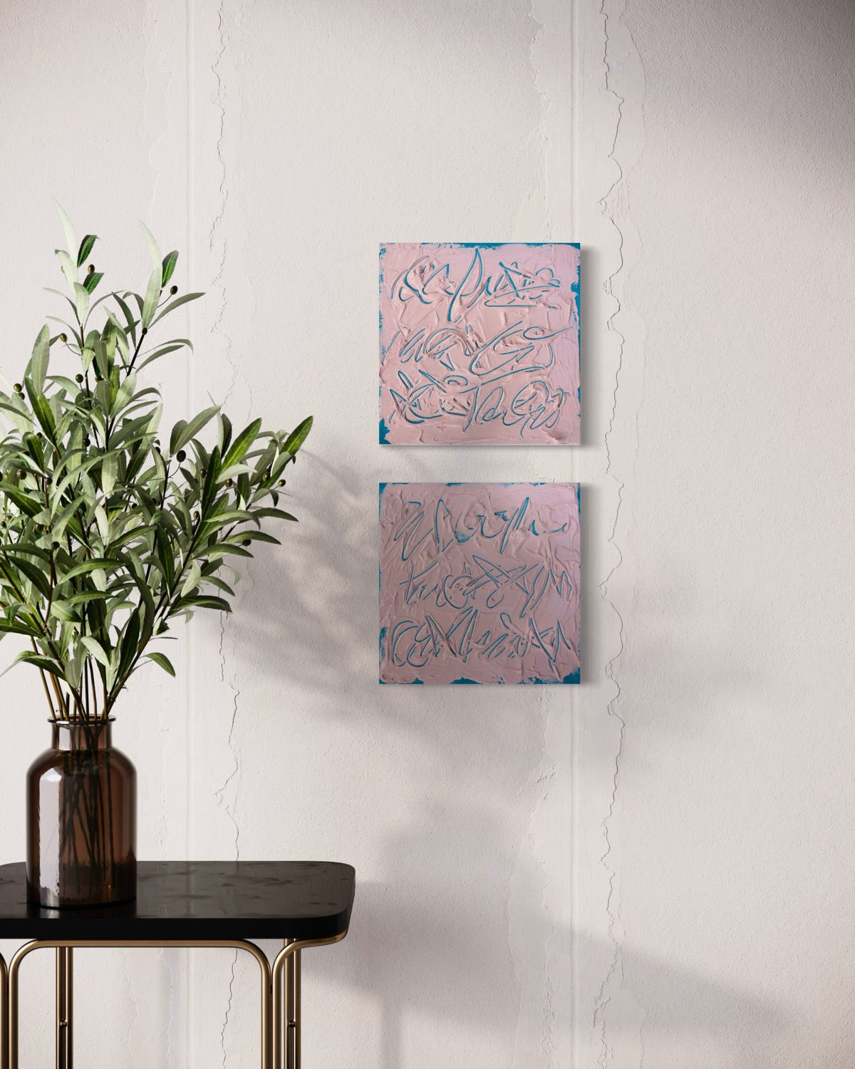 Two square abstract texted based paintings with pink backgrounds and teal line work hang on a white wall above a black side table with a green plant in a brown vase.