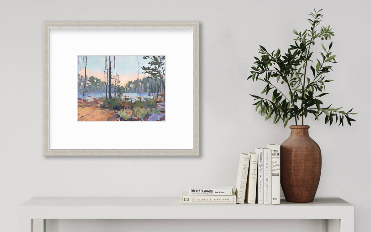 A light blue, green, brown and purple abstract landscape print in a silver frame with a mat hangs on a white wall above a white console table with a stack of books and a greenery filled terracotta vase.