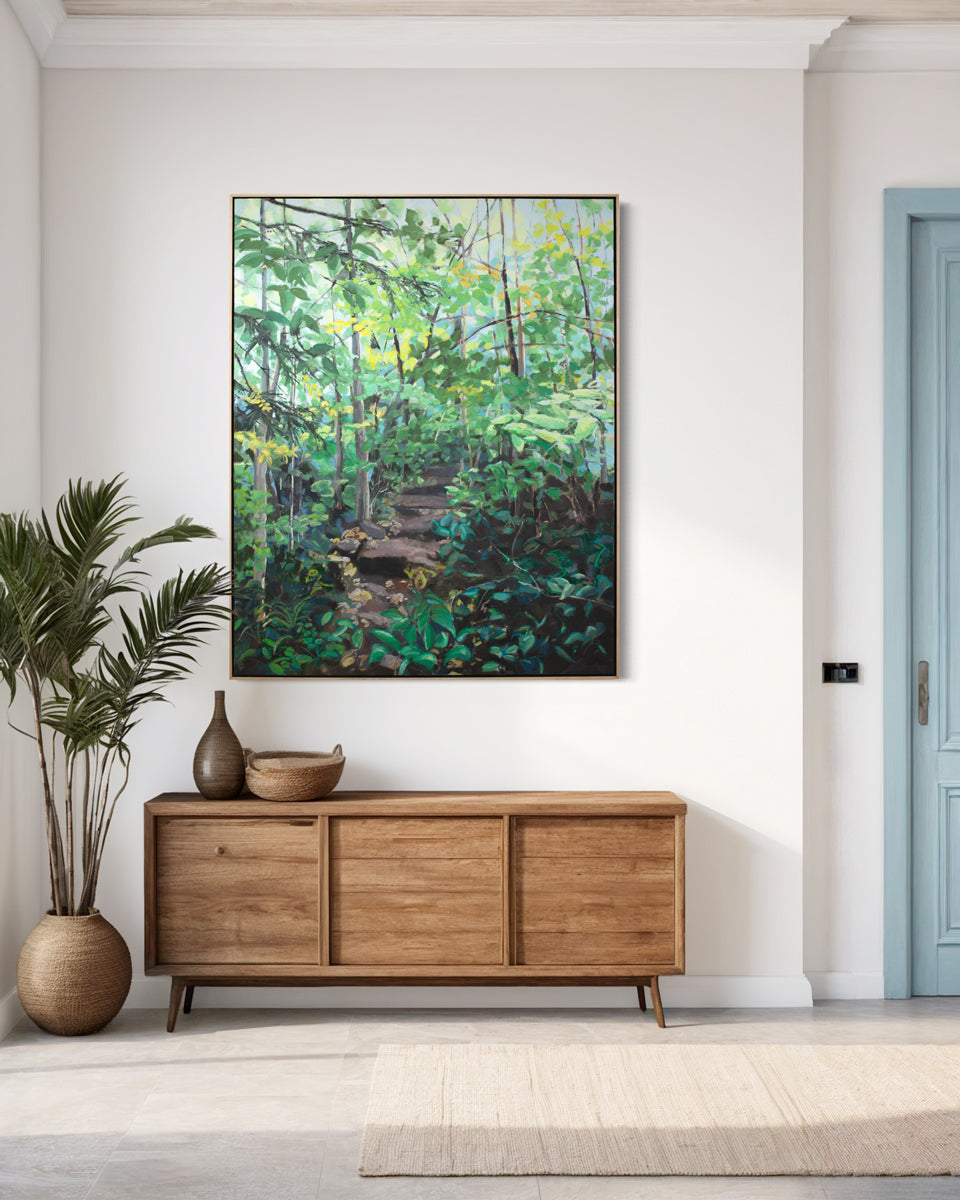 A large framed green painting of a forest hangs on a white wall above a wooden console table, potted floorplant, and next to a light blue door.