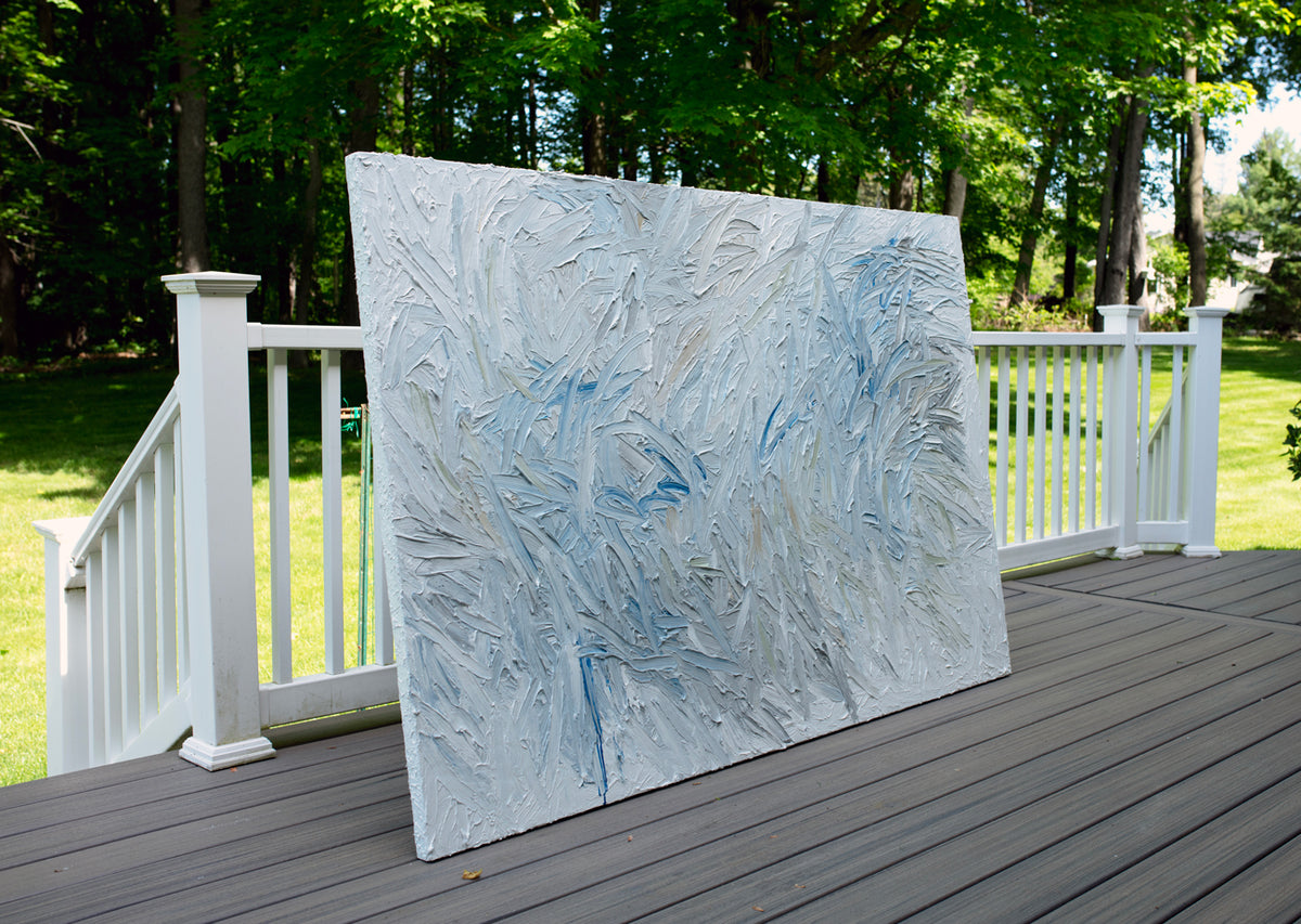 Abstract painting on a wooden deck with a white railing and greenery in the background.