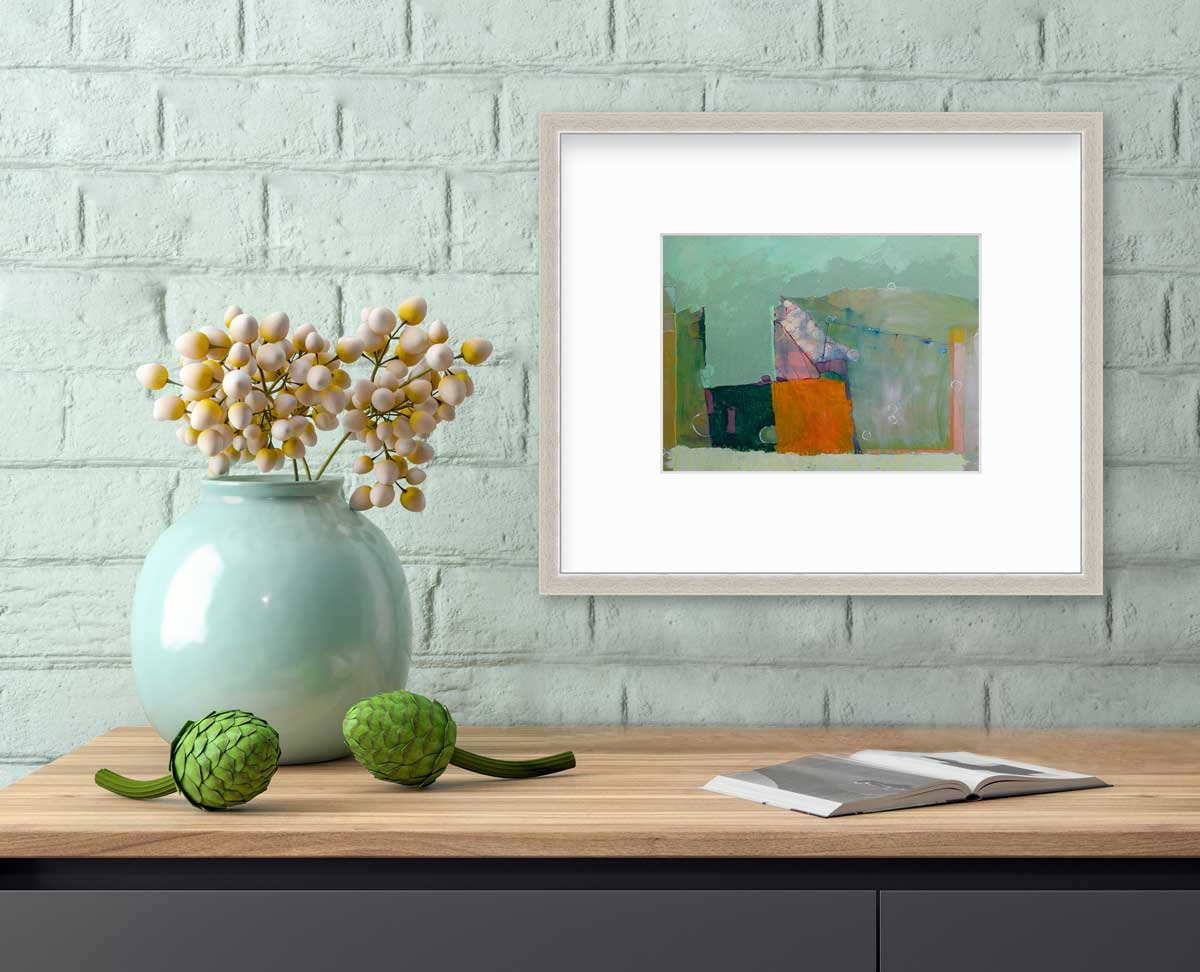 A green abstract print by Christine Averill-Green in a silver frame with a mat hangs on a light teal brick wall above a wood grain surface with an open book, two green succulents and a teal vase with pink and yellow buds.
