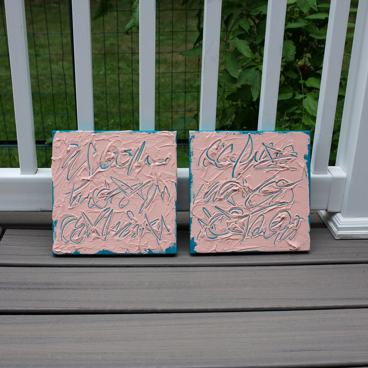 Two square pink textured abstract text based paintings with teal abstract brushstrokes, displayed side-by-side on a gray composite deck in front of a white railing with green foliage in the background.