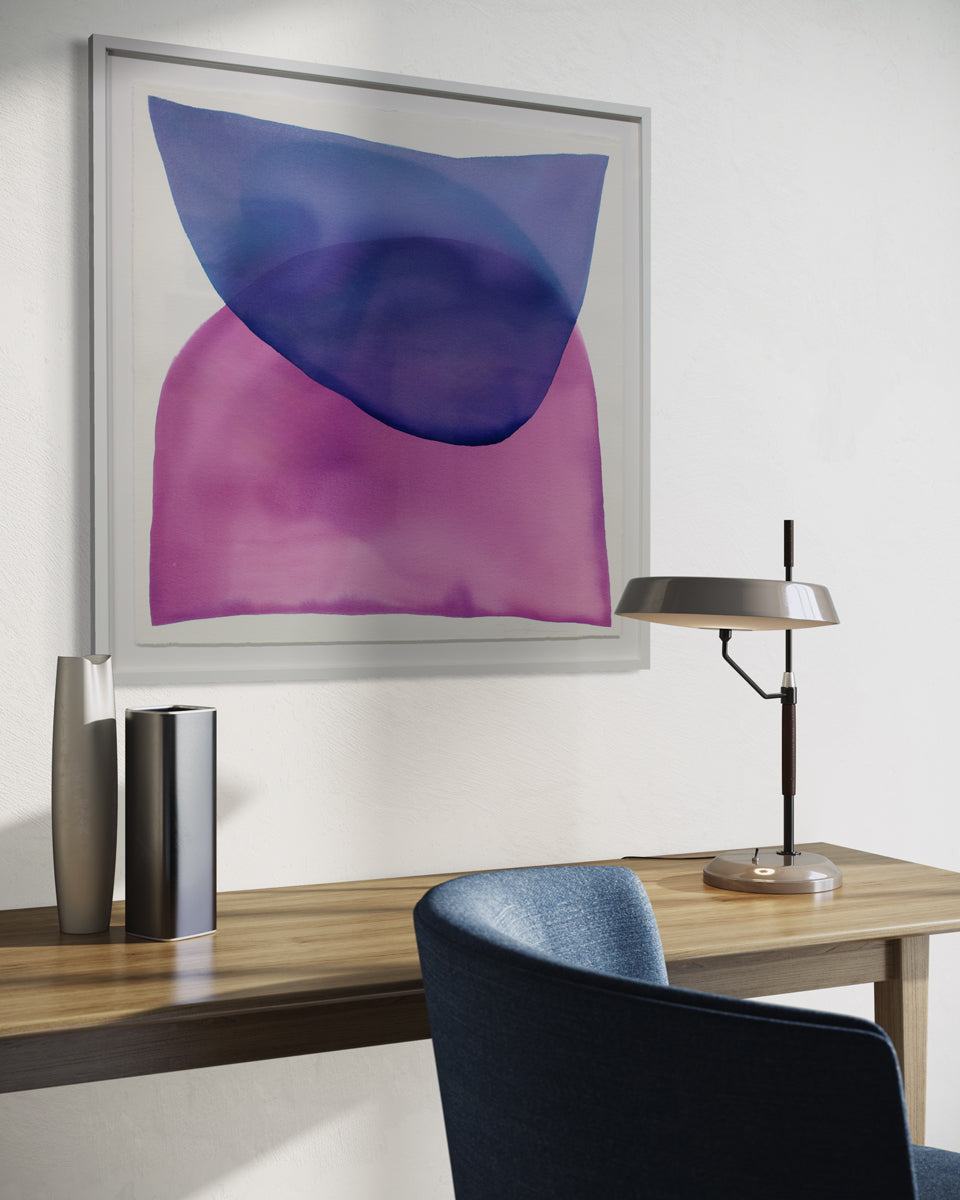 A blue and magenta minimalist abstract watercolor painting framed in a white frame hangs on a white wall above a simple wood desk with a table lamp, simple table vases and a blue upholstered desk chair.