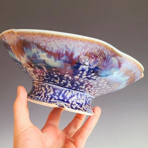A hand holds a multicolored ceramic serving bowl in front of a grey gradient backdrop.