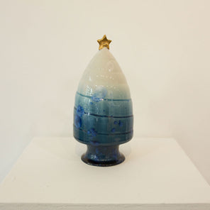 A sculpture of a tree that fades from dark blue at the base up to white at the top of the tree with a gold star on top rests on a white pedestal in front of a white wall. 