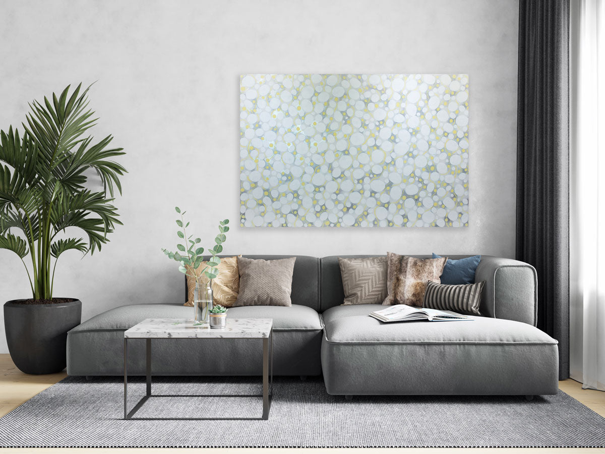 An abstract white, yellow, and grey painting hangs on a light grey wall above a modern grey sofa and coffee table, and between a large potted floor plant and grey curtains hanging in a window.
