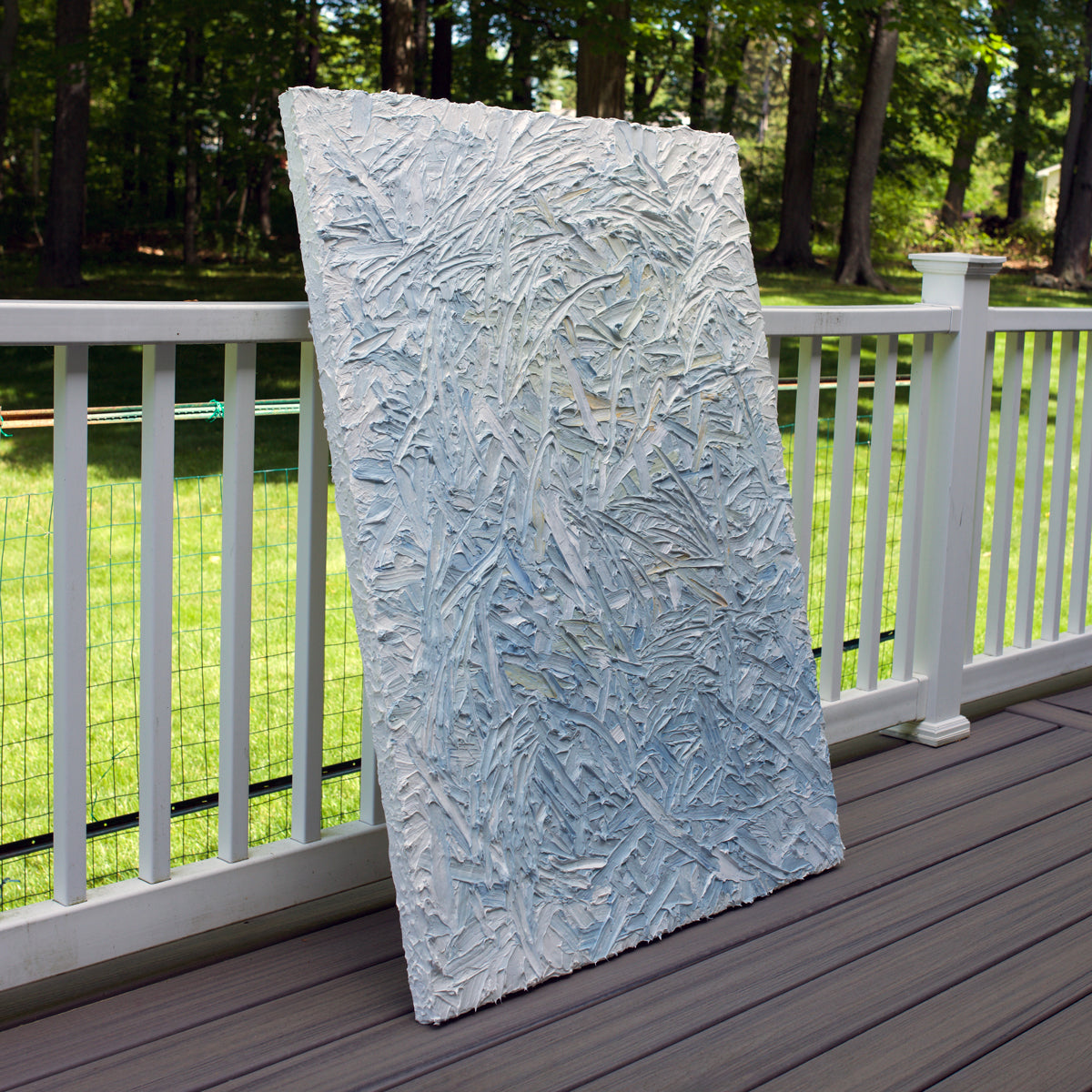 The sideview of an original abstract heavily textured painting on a wooden deck with a green lawn and trees in the background
