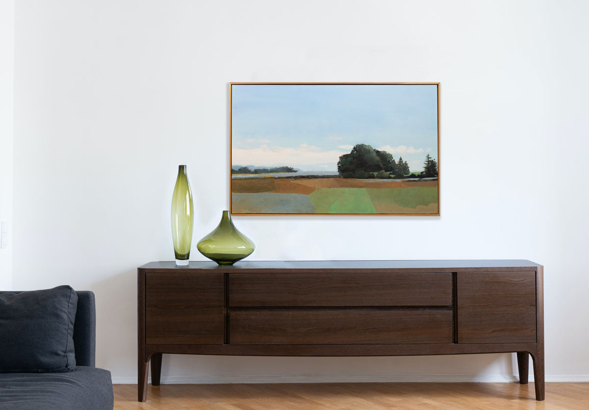 An earth-toned landscape painting hangs on a white wall above a dark wood console table and green glass table decor.