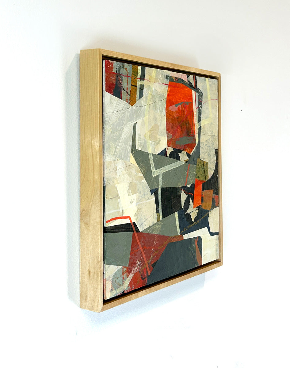 A framed abstract collage artwork featuring geometric shapes in shades of red, beige, and green, with a white background hanging on a white wall, side view.