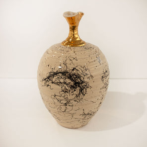 A glazed cream-colored vase with black finish and gold neck rests on a white surface in front of a white wall. 