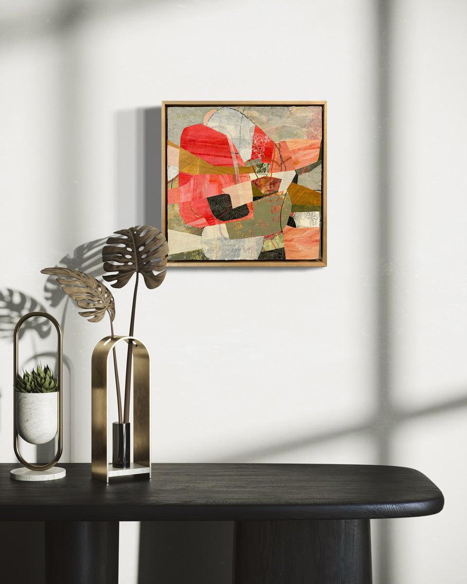 A framed abstract collage artwork featuring geometric shapes in shades of red, beige, and green, with a white background hanging on a white wall.