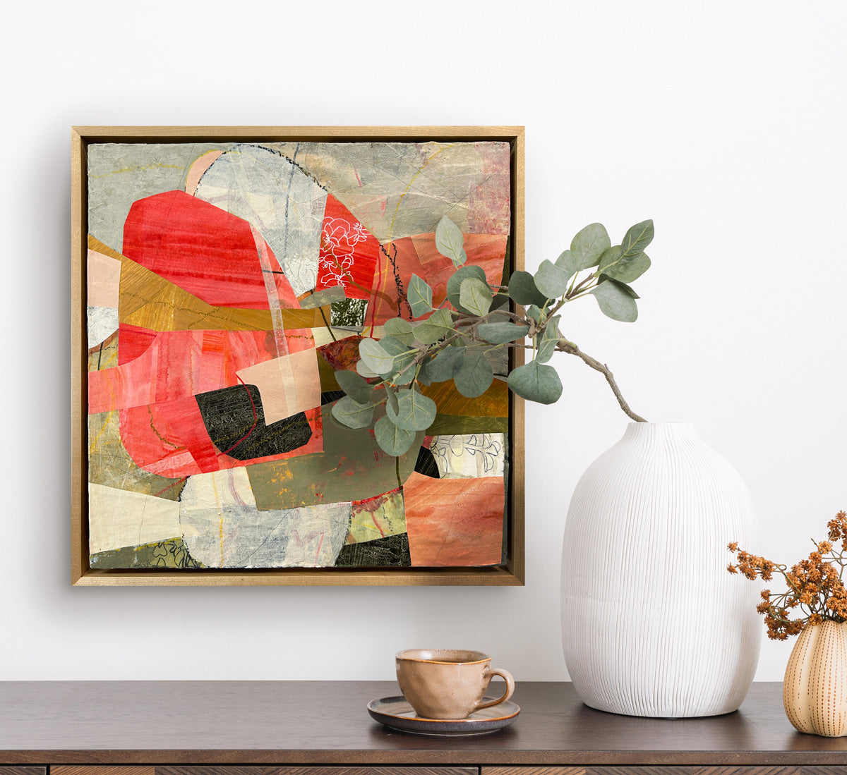 A framed abstract collage artwork featuring geometric shapes in shades of red, beige, and green, with a white background hanging on a white wall.