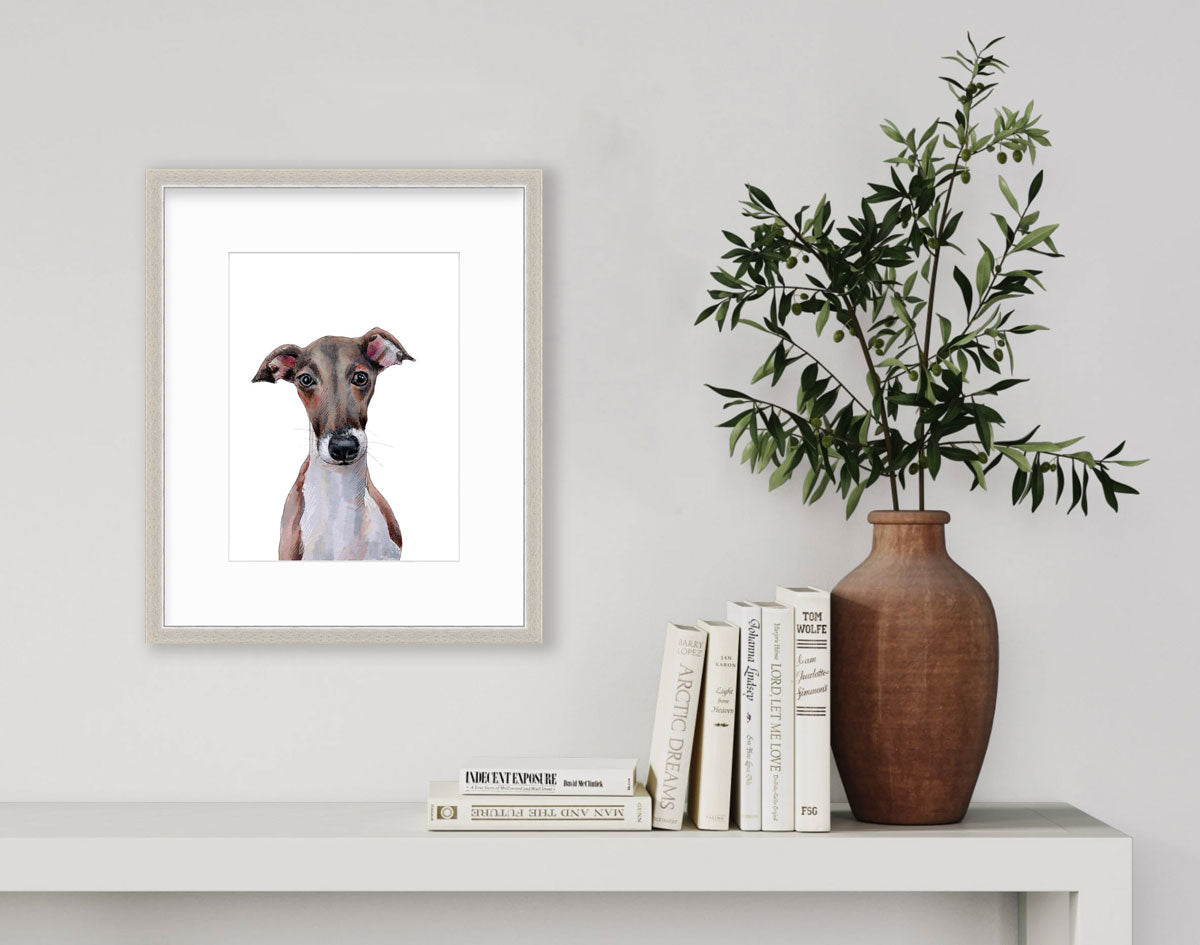 An illustration of an Italian Greyhound dog framed in a silver frame hangs on a light wall above a modern white console table with books and a plant in a brown ceramic vase.