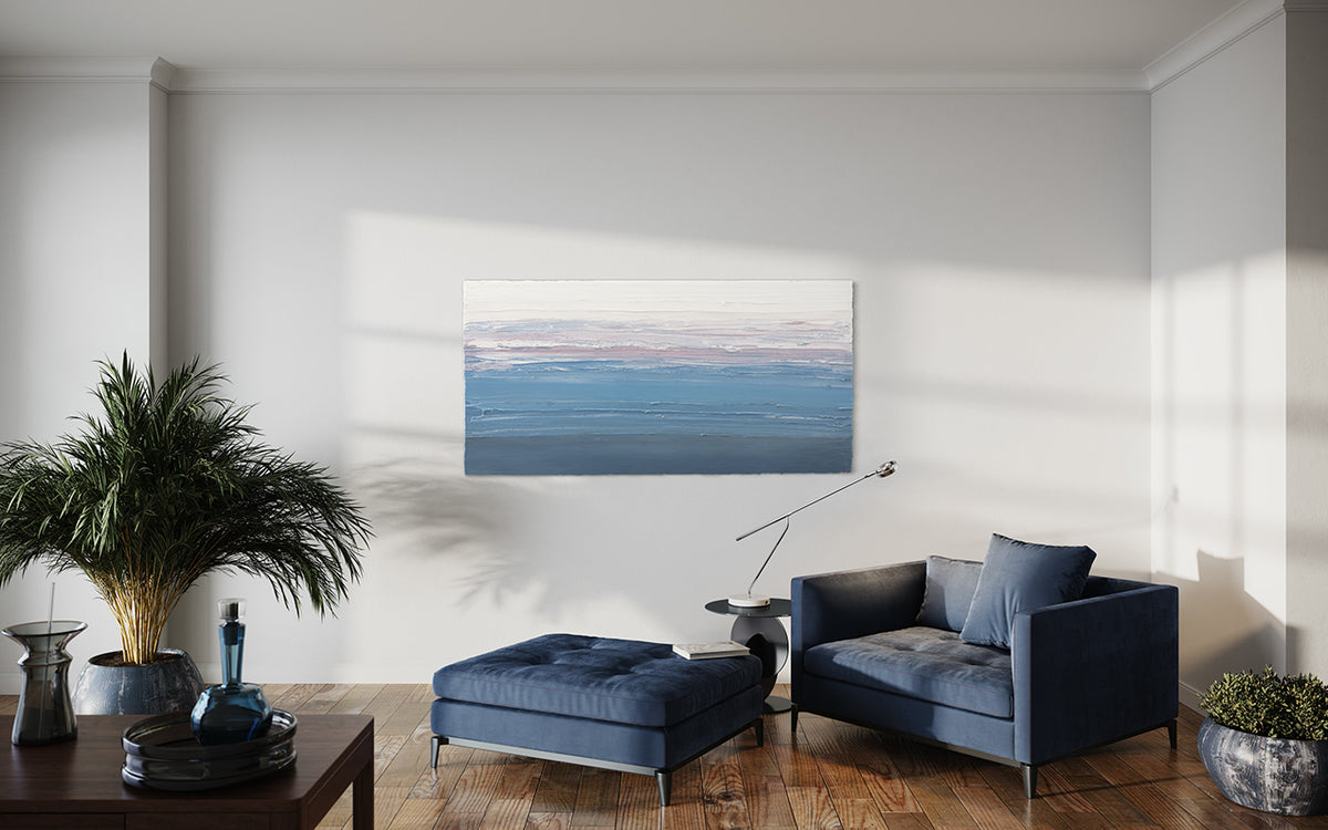 A blue, lavender and white thickly textured abstract painting by Teodora Guererra on a white wall in natural light hangs over a blue oversized chair with ottoman. A contemporary end table is to the side of the chair with a modern light. Also, two plants are on either side and a coffee table is to the left with one blue bottle and other on it.