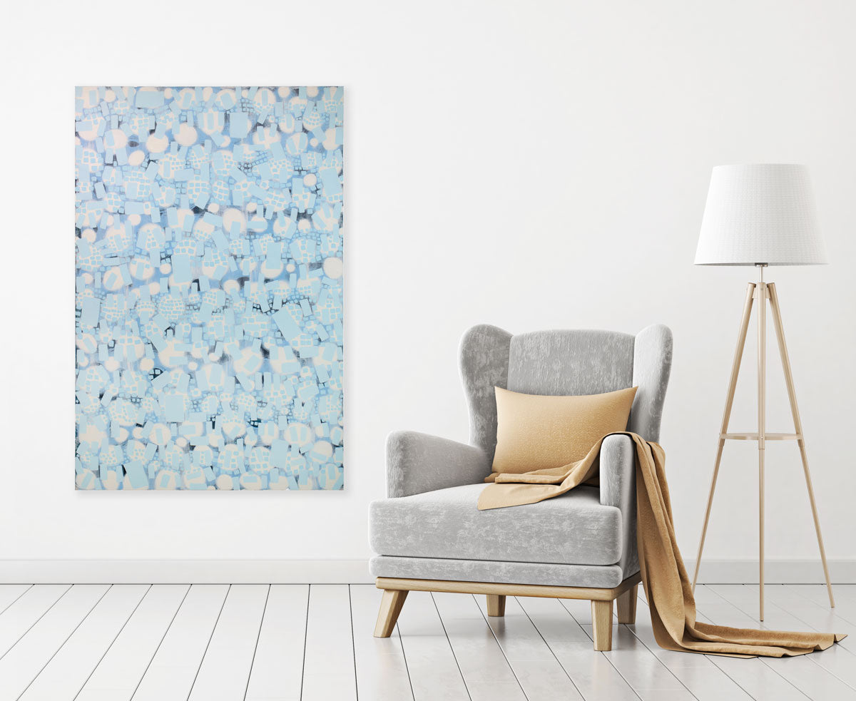 A white and blue abstract painting hangs on a white wall next to a grey armchair with a muted yellow throw and pillow, and a standing floor lamp.