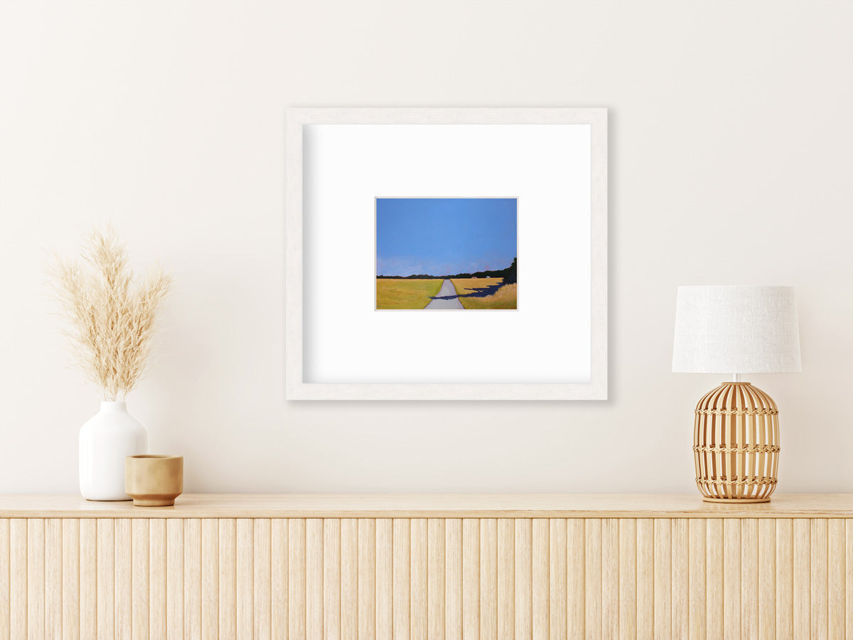 A blue and yellow-green abstract landscape print, framed in a white wood frame hangs on a light wall above a light wooden console table with light table decor.
