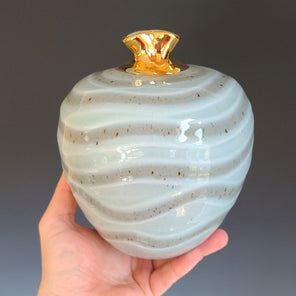 A hand holds a light green and sandy beige ceramic vessel with a flared gold neck in front of a grey gradient background. 