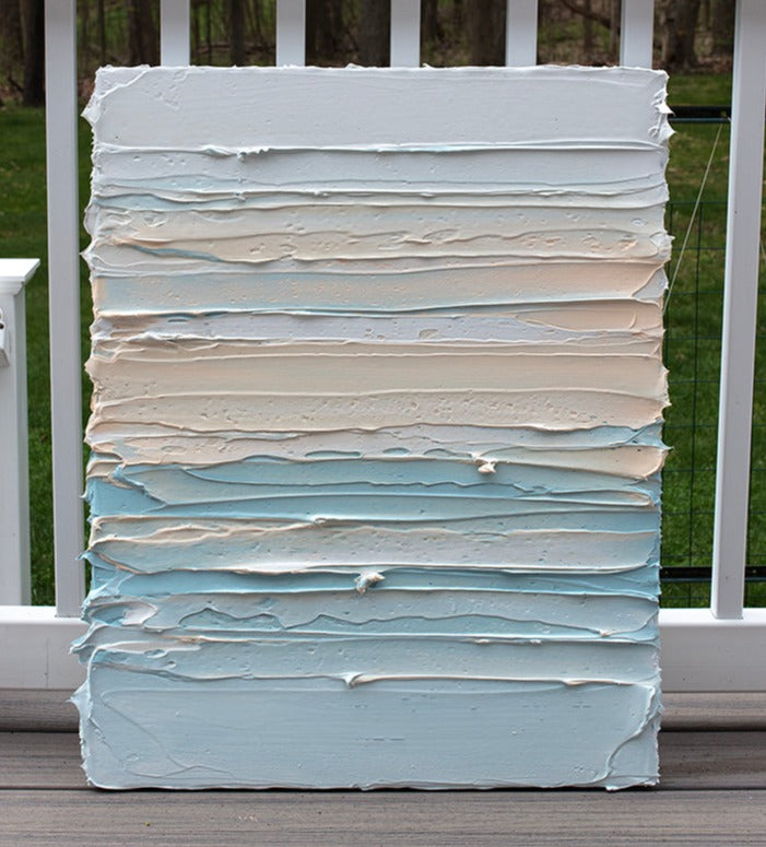 An abstract painting with heavily textured white, sherbet orange and teal blue green paint sitting on the artists deck outside.