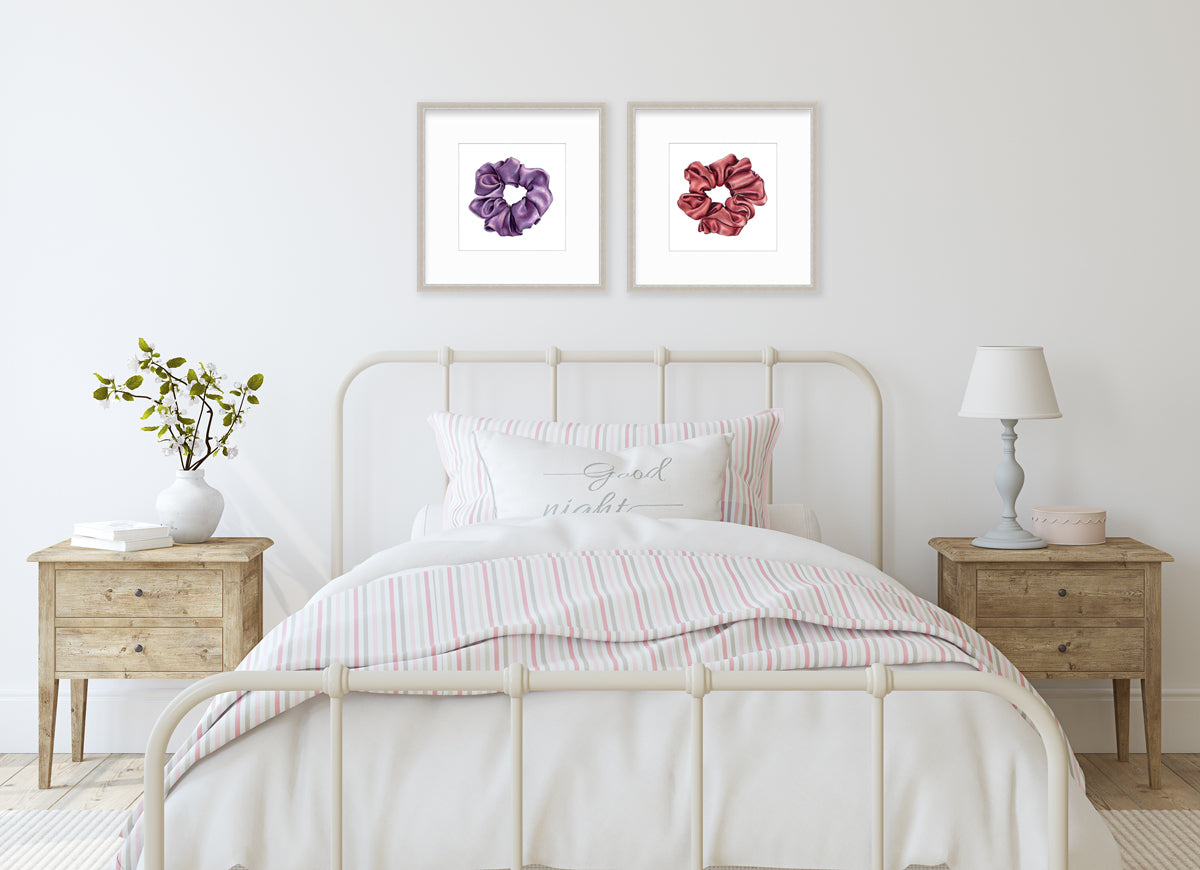Two art prints of hair scrunchies framed in silver frames hang on a light wall above a pink, grey, and white striped twin bed and light wood end tables.