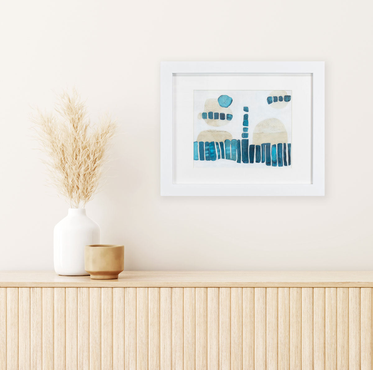 A blue, beige, and white painting in a white frame hangs on a light wall above a wooden console table with white and beige table decor.