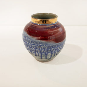 A red and blue ceramic vase with a round gold neck rests on a white surface in front of a white wall. 