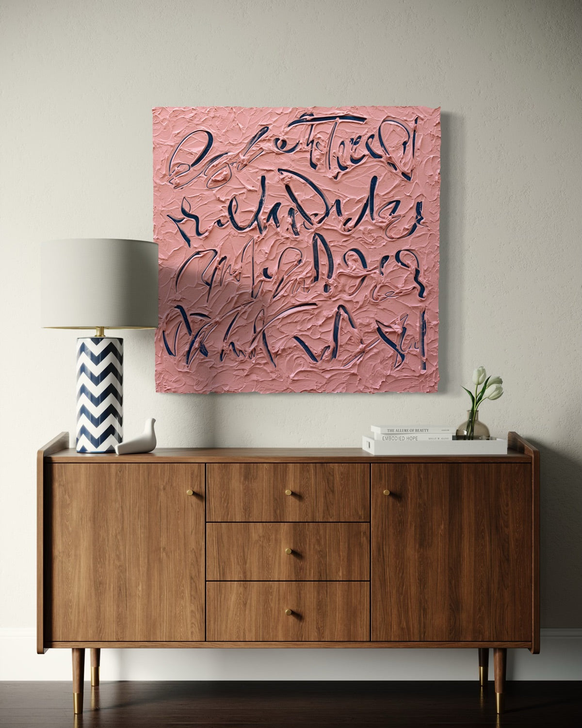A square text based pink textured painting with dark blue abstract calligraphy hangs on a light gray wall above a mid-century modern wooden sideboard with a lamp and small plant.