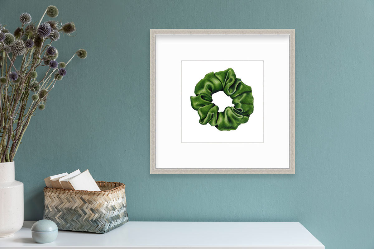 A drawing of a green hair scrunchie framed in a silver frame hangs on a teal wall above a white table with boho decor.