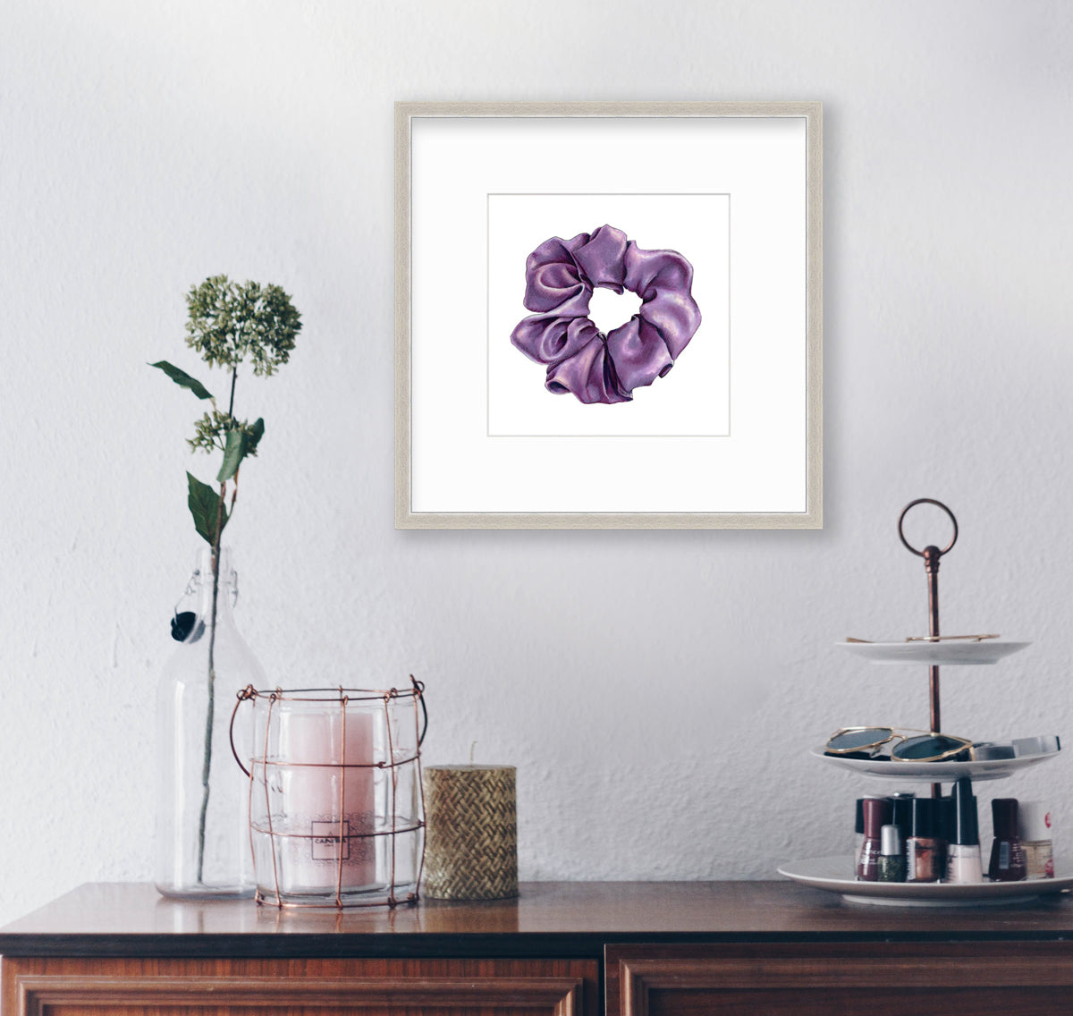 A drawing of a purple hair scrunchie, framed in a silver frame hangs on a white wall above a dresser with table decor and a stand with nail polish and sunglasses.