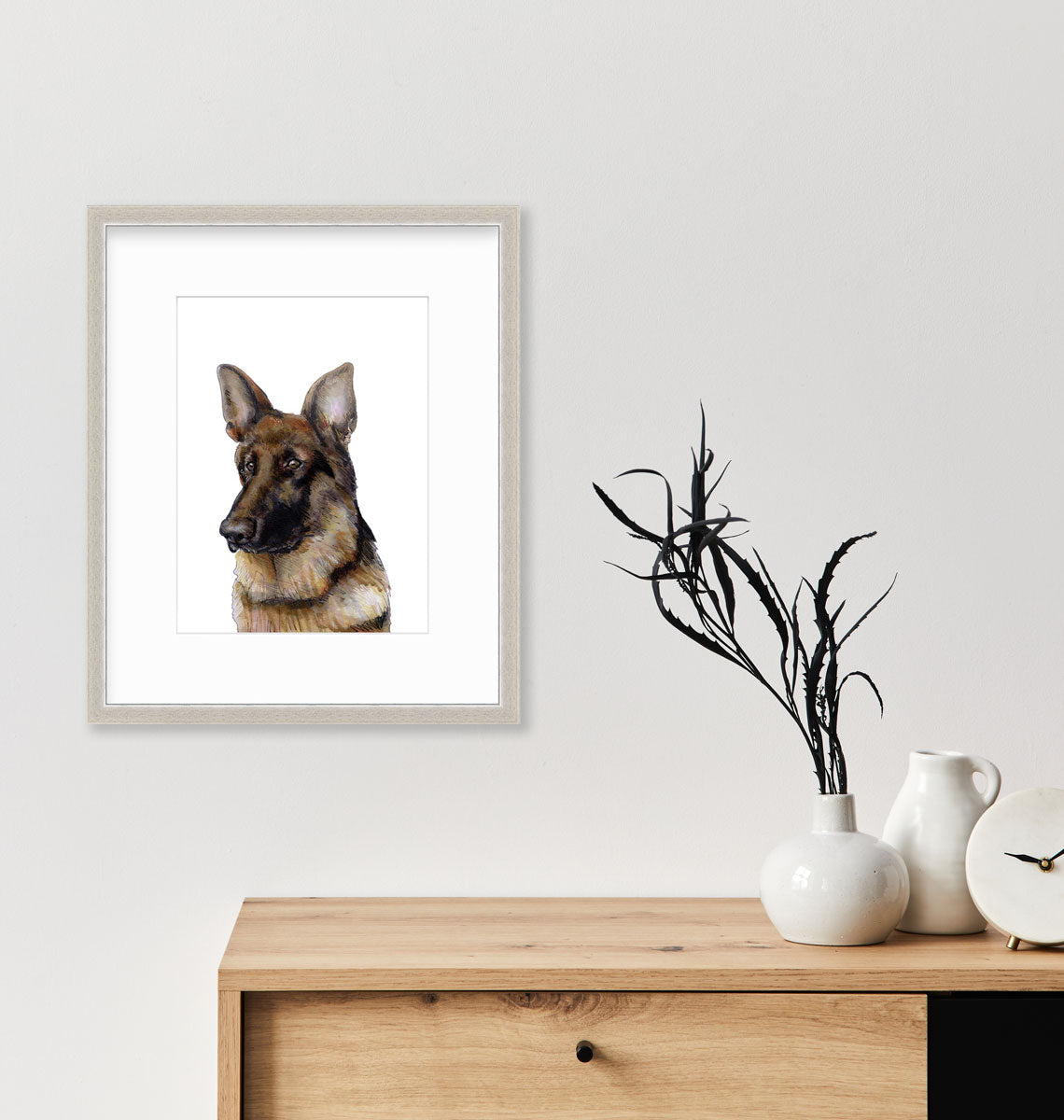 An illustration of a German Shepherd dog framed in a silver frame hangs on a white wall above a wooden console able with white modern table decor.