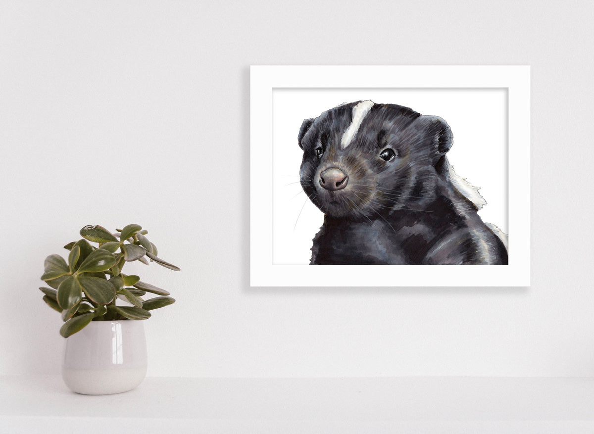 A framed illustration of a skunk hangs on a light wall above a table with a small potted plant.