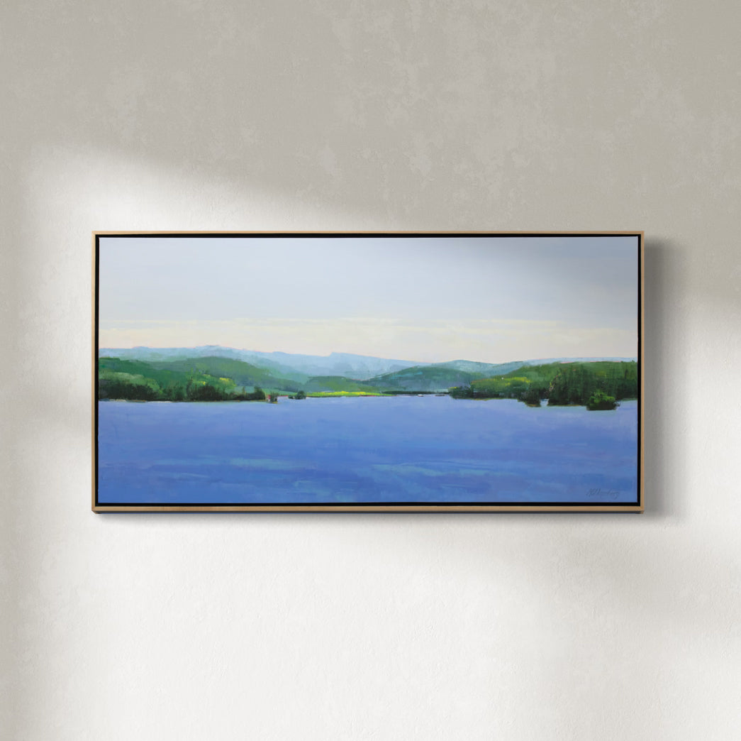 A blue and green landscape painting framed in a light wood frame hangs on a white wall.