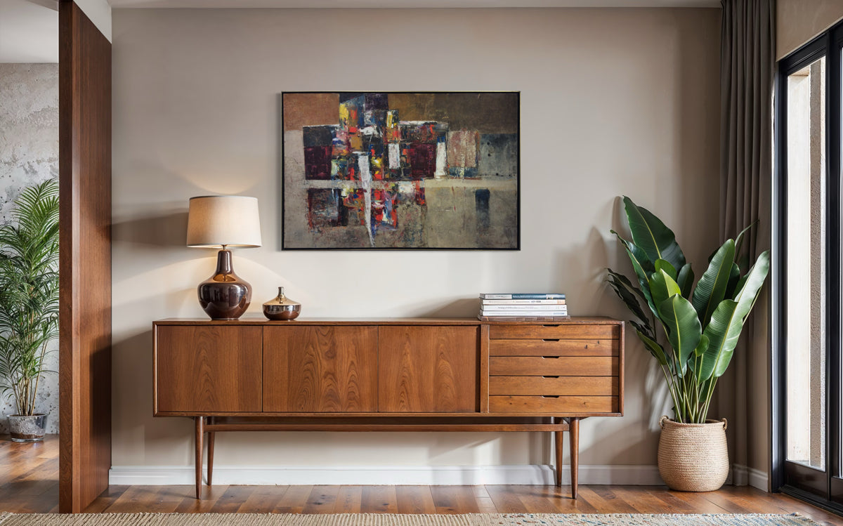 Wooden sideboard with abstract painting on the wall in a room with plants and a lamp.