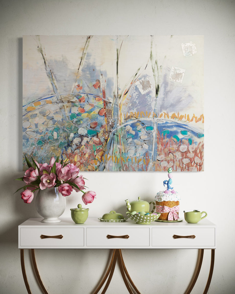A multicolored abstract painting hangs on a white wall above a white credenza with flowers and decorative items.