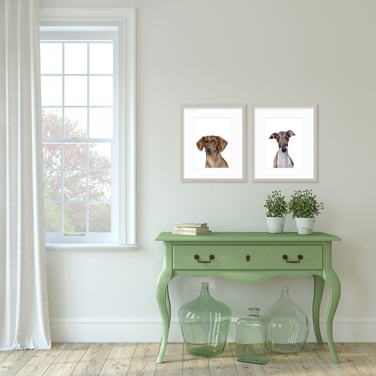 Two illustration prints of dogs framed in silver frames hang on a light grey wall above a green console table with glass vases, books, and potted plants. A large white window is to the left.