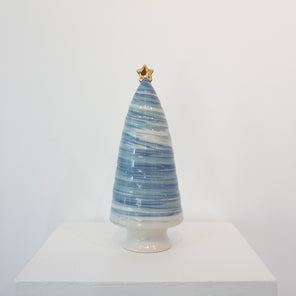 A blue and white ceramic sculpture of a tree with a gold star on top sits on a white pedestal in front of a white wall. 