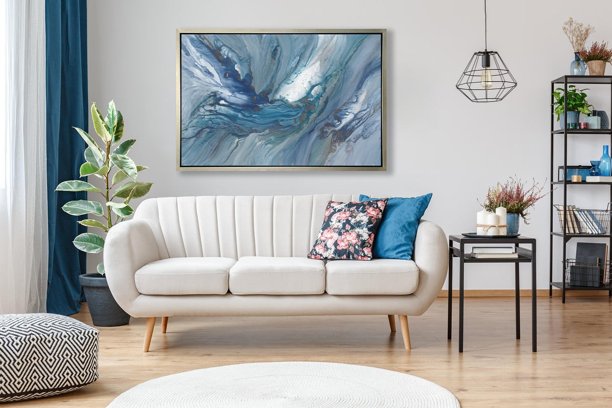 A blue, teal and white abstract print in a silver floater frame hangs on white wall above a white sofa with printed pillows, a black side table with accessories and a potted floor plant.